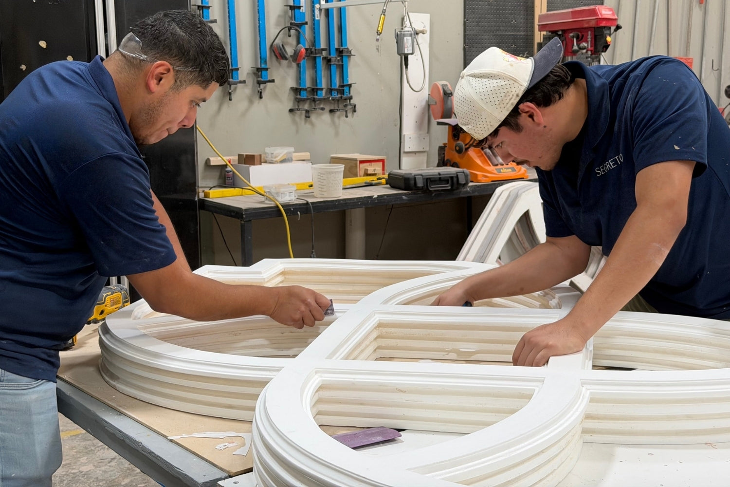 Two men working together in a workshop, assembling a large white composite wood frame. One man wears a navy blue t-shirt and jeans, while the other wears a navy blue t-shirt, a white cap, and light jeans.