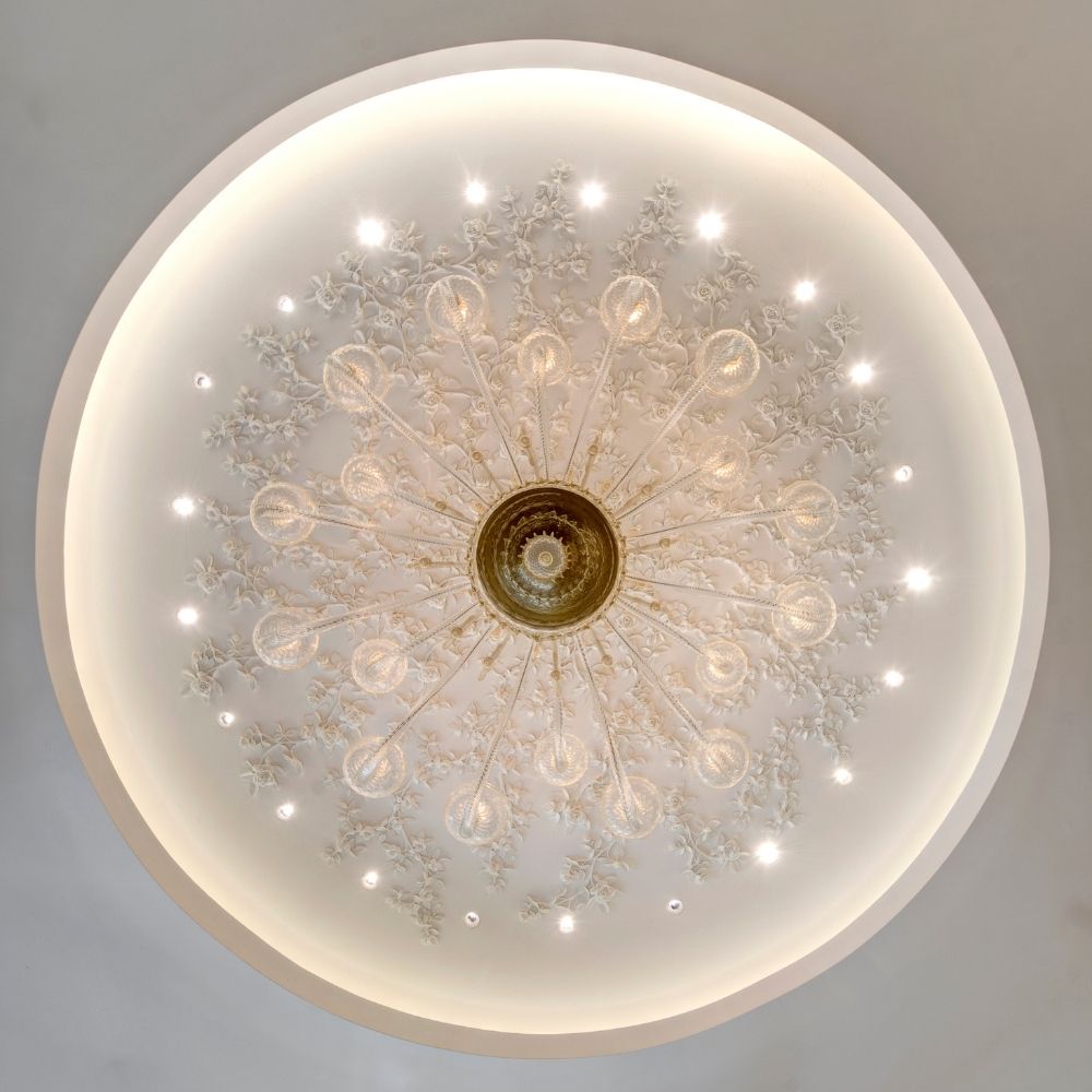 A large sculptural plaster ceiling chandelier with intricate floral detailing and multiple illuminated light points, shown from a top-down perspective against a light gray ceiling.