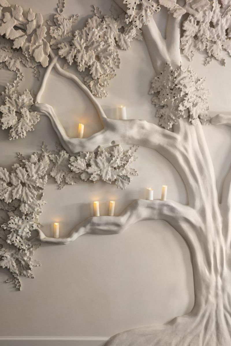 A close-up of a sculptural white tree branch and leaf wall piece with several small lit candles nestled in the branches against a light neutral by background by Segreto Finishes.